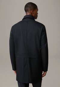 Zwarte jas met een hoge kraag, een textuurstof en zichtbare naden. Heeft een aansluitende pasvorm, een split aan de achterkant en zijzakken.