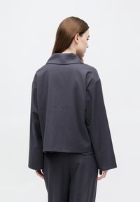 Sivá krátka bunda s ostrým golierom, spustenými ramenami a dlhými rukávmi, vyrobená z hladkej látky. Dizajn je minimalistický.