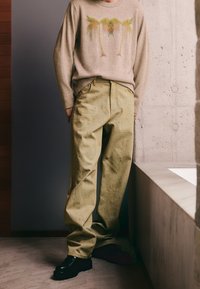 Pull beige avec un motif de palmier, associé à un pantalon ample vert clair. Des chaussures noires épaisses complètent la tenue.