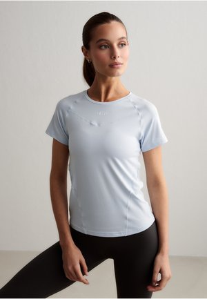 Vrouw in een lichtblauw sportshirt met korte mouwen en zwarte leggings, poserend met één gebogen been, lichtjes opzij kijkend tegen een effen achtergrond.