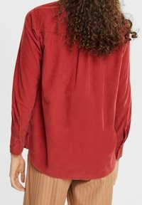 Chemise rouge à manches longues en tissu à texture douce, avec un poignet boutonné et un ourlet arrondi. Vue de dos.