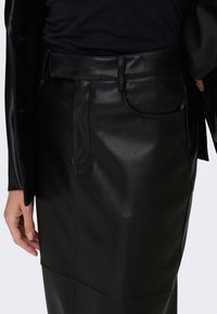 Gros plan d'une personne portant une jupe en cuir noire et une veste assortie, avec une main reposant à côté.