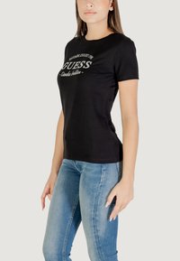 T-shirt nera in cotone con maniche corte, con un logo bianco centrale. Abbinata a jeans di denim blu chiaro. Design semplice e aderente.