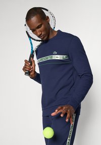 Tamsiai mėlynas sportinis megztinis su apskritu kaklu, su Lacoste logotipu ir dryžuota dizaino. Laikantis teniso raketę, krentantis neoninės žalios spalvos kamuolys.