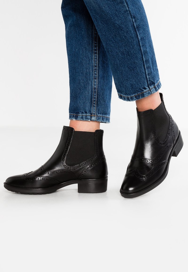 Botas de cuero negro hasta el tobillo con paneles laterales elásticos, detalles decorativos de broguing y un tacón bajo tipo bloque. Combinadas con jeans de mezclilla azul doblados.