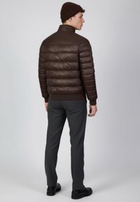 Chaqueta acolchada marrón con textura brillante y costuras segmentadas horizontales. Combinada con pantalones gris oscuro con estampado y un gorro de punto a juego.