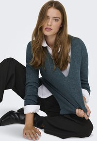 Tiefblaue Strickjacke mit tiefem V-Ausschnitt, kombiniert mit einem weißen Hemd mit Kragen. Modell sitzt in schwarzer Hose und Stiefeletten und zeigt die Textur des Stoffes.