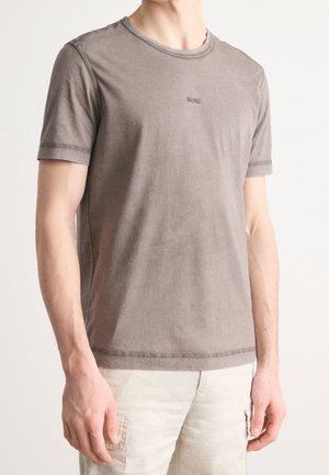 Mann trägt ein verblasstes braunes BOSS-T-Shirt mit kurzen Ärmeln und helle beige Cargo-Hosen, steht vor einem schlichten Hintergrund.