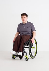 Μαύρο αναπηρικό καροτσάκι με πράσινες λεπτομέρειες; γκρι μπλουζάκι; καφέ παντελόνι φαρδύ; λευκά αθλητικά παπούτσια; κοντά μαύρα μαλλιά; απλό λευκό φόντο.