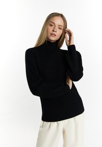 Maglione a coste nero con colletto alto; presenta maniche lunghe e una silhouette aderente. Indossato con pantaloni color crema.