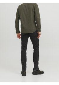 Camicia verde oliva a maniche lunghe con polsini a bottoni, abbinata a jeans neri e stivali neri alla caviglia, mostrata di spalle su uno sfondo neutro.