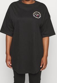 Czarny, oversize t-shirt noszony przez osobę, z napisem "Tommy Jeans NYC" w okrągłym wzorze na lewej stronie klatki piersiowej.