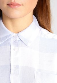 Lys blå og hvidternet skjorte med knap-ned krave, der har et tekstureret stof og en enkelt lomme på venstre side.