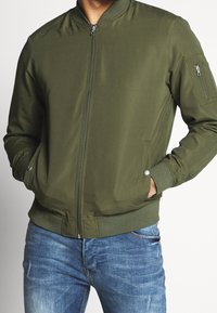 Man in een olijfgroene bomberjack met handen in de voorzakken, gecombineerd met vervaagde blauwe jeans tegen een effen achtergrond.