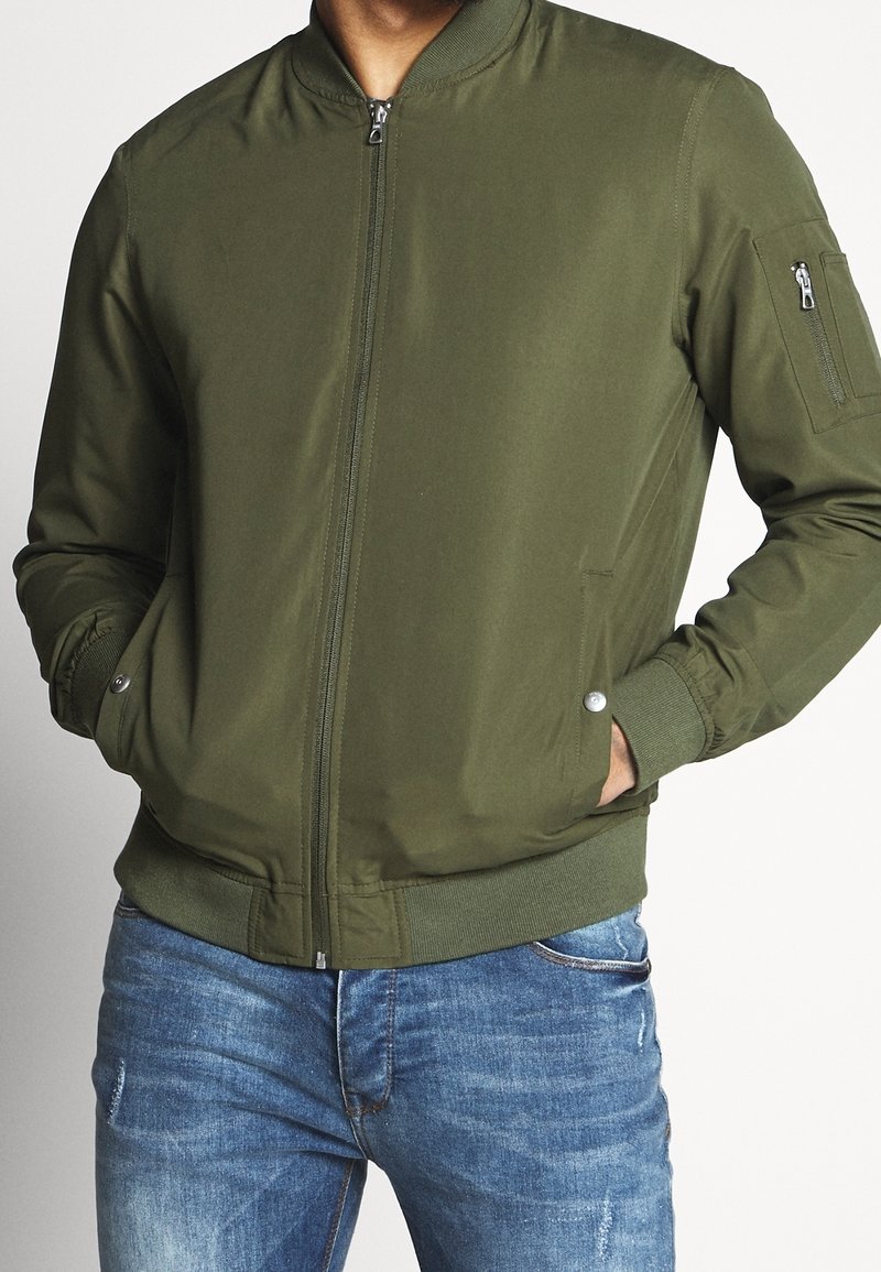 Man in een olijfgroene bomberjack met handen in de voorzakken, gecombineerd met vervaagde blauwe jeans tegen een effen achtergrond.