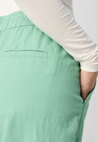 Pantalons verts clairs, coupe décontractée, en tissu texturé, dotés d'une poche latérale et d'un élastique à la taille, associés à un haut blanc à manches longues.