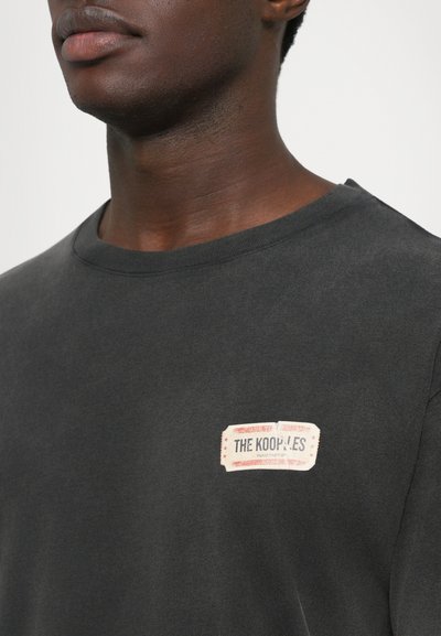 Homem a usar uma t-shirt crewneck cinza escuro com um pequeno retângulo de logotipo "THE KOOPLES" no lado esquerdo do peito.