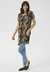 Tunique imprimée léopard en beige, marron et noir, avec des manches courtes et un col froncé, associée à un jean bleu et des ballerines noires.