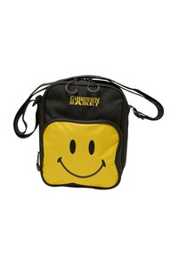 Sort taske med design af gult smiley-ansigt. Har justerbar rem, lynlås-lukning og "CHINATOWN MARKET" logo i gult øverst.