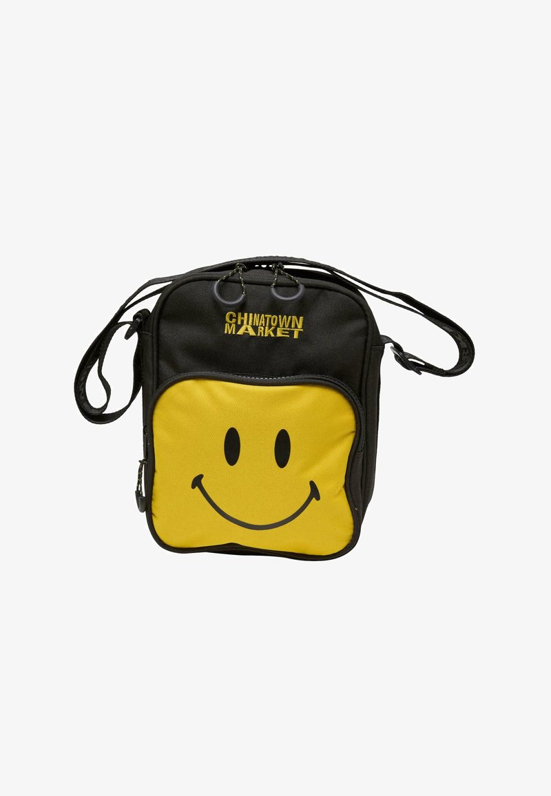 Sort taske med design af gult smiley-ansigt. Har justerbar rem, lynlås-lukning og "CHINATOWN MARKET" logo i gult øverst.