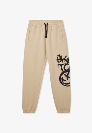 Beżowe dresy wykonane z miękkiego materiału, z elastycznym paskiem w talii z sznurkiem, bocznymi kieszeniami oraz czarnym graffiti na udzie.