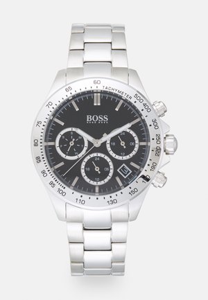 Montre-bracelet Hugo Boss argentée avec cadran noir, trois sous-cadrans pour le jour, les 24 heures et les secondes, lunette tachymètre et fenêtre de date à 4 heures.