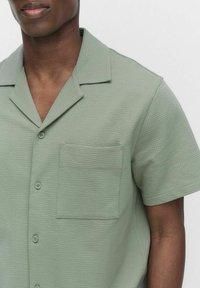 Chemise verte à manches courtes en tissu texturé, avec un col en V, une poche poitrine et une fermeture à bouton.