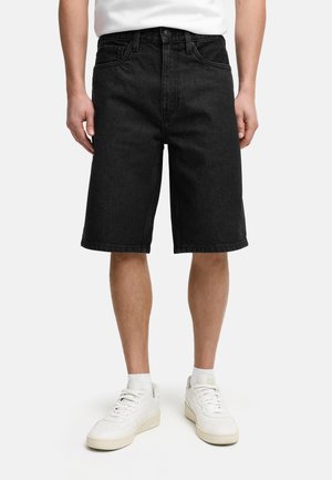 Mand iført sorte denim knælange shorts, hvide sneakers og en hvid t-shirt, stående mod en lys baggrund.