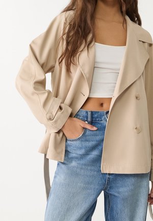 Frau trägt beige übergroße Jacke, weißes kurzes Top und hellblaue, hoch taillierte Jeans, eine Hand in der Tasche.