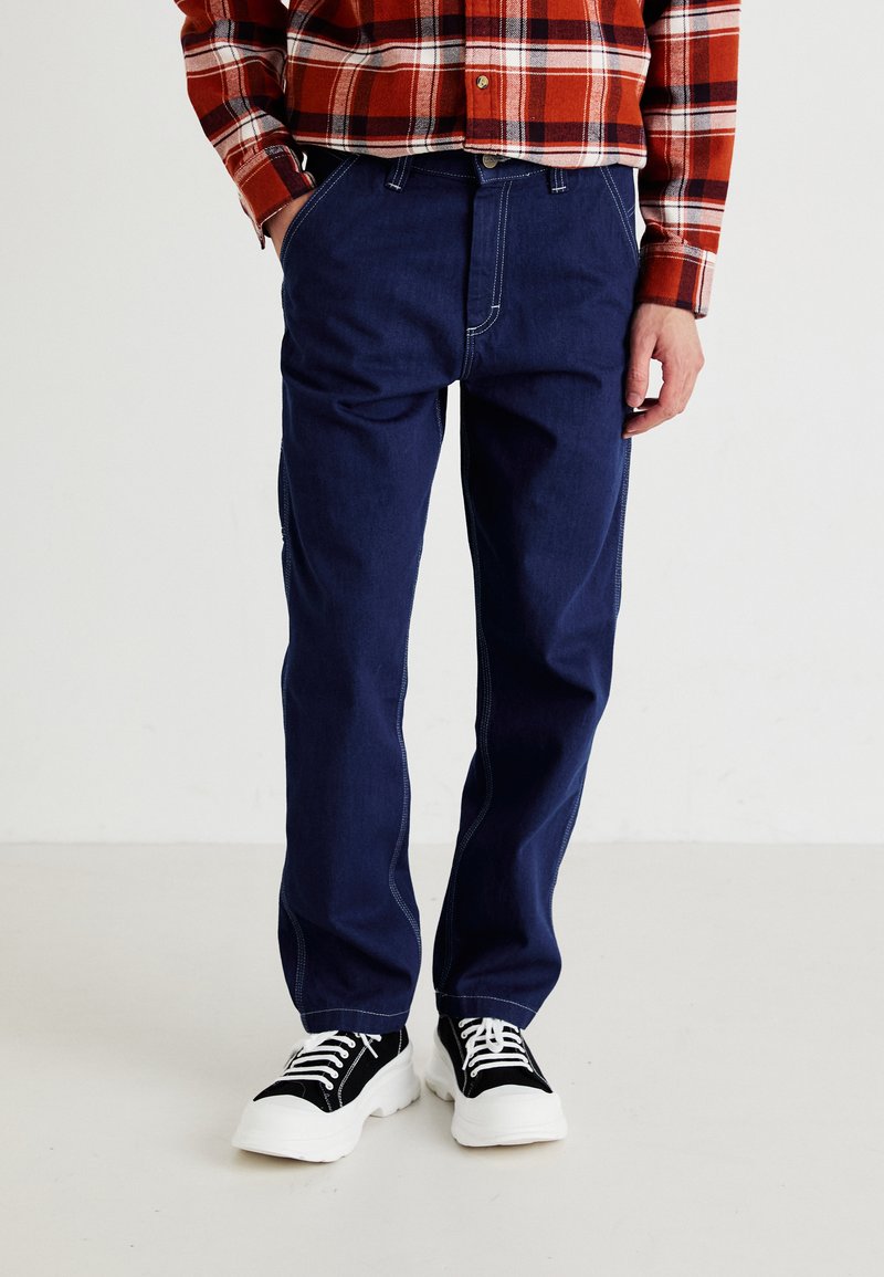 Personne portant un jean bleu foncé, des baskets épaisses noires et blanches, et une chemise à carreaux rouges, les mains dans les poches, debout sur un sol uni.