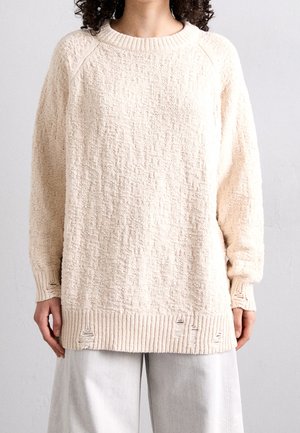 Frau trägt locker sitzenden, strukturreichen cremefarbenen Pullover mit ausgefranstem Saum und Ärmeln über hellgrauer Hose, steht vor neutralem Hintergrund.