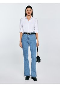 Žena stojící, oblečená v bílé košili na knoflíky, světle modrých džínsách s vysokým pasem, černém pásku, černých botách a držící černou kabelku se zlatým řetízkem.