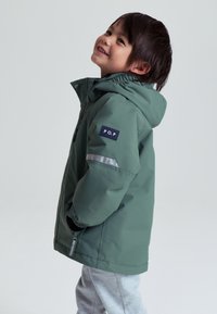 Grøn vinterjakke med hætte, der har en frontlynlås og trykknaplukning, reflekterende stribe på ærmet og et logo-patch på armen.