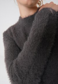 Grauer flauschiger Pullover mit hohem Kragen und langen Ärmeln, der eine weiche Textur und sichtbare Strickmuster aufweist.
