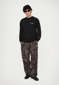 Zwart shirt met lange mouwen met de tekst "No Problemo", gecombineerd met losse, bruine broek met camo-patroon. Zwarte zonnebril, zwarte schoenen.