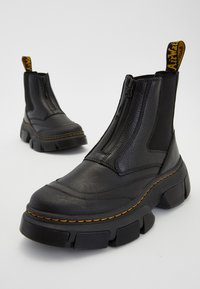 Paire de bottes épaisses en cuir noir avec fermetures éclair frontales, coutures jaunes et semelles épaisses texturées sur un fond clair uni.