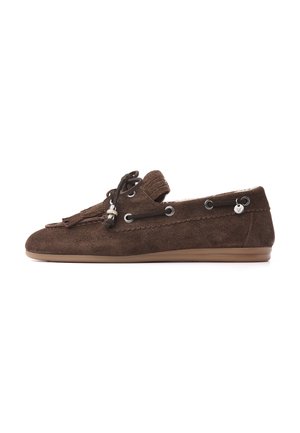 Bruine suède loafer met kwastjes, zwarte veterdetail door metalen oogjes en een lage rubberen zool op een witte achtergrond.