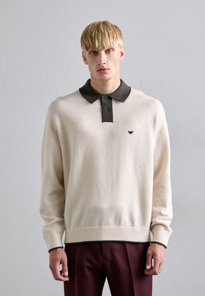 Jeune homme portant un pull beige avec un col et des poignets foncés, associé à un pantalon rouge foncé, se tenant contre un mur gris clair uni.
