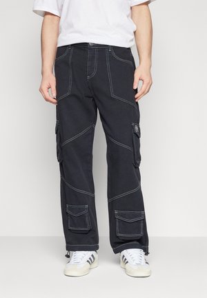 Mann trägt schwarze Cargohose mit großen Seitentaschen und weißen Sneakers, stehend vor hellem, einfarbigem Hintergrund.