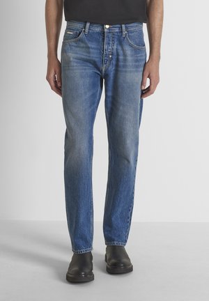Uomo che indossa jeans blu classici a gamba dritta e stivali neri slip-on, in piedi davanti a uno sfondo grigio chiaro uniforme.