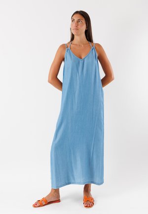 Abito maxi senza maniche di un blu chiaro, realizzato in tessuto denim. Disegno a V con spalline sottili e vestibilità rilassata, completato da sandali arancioni.