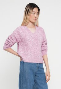 Helles rosa V-Ausschnitt-Pullover mit einem strukturierten Strickmuster und überschnittenen Schultern, kombiniert mit hochgeschnittenen blauen Jeans.