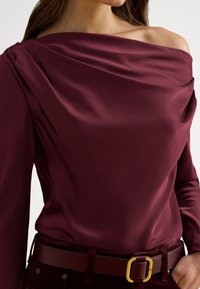 Bordový saténový top s drapírovaným výstrihom, dlhými rukávmi a jemnými detailmi na ramenách. Doplnýva ho zladený opasok so zlatou prackou.