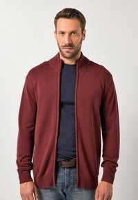 Bordó pletený kardigan s vysokým límcem, vroubkovanými manžetami a lemem, nošený přes námořnickou košili, s předním zipovým zapínáním.