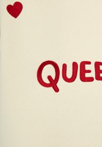 Tessuto bianco con ricami rossi. Mostra la parola "QUEEN" in lettere in grassetto con un accento a forma di cuore rosso nell'angolo in alto a sinistra.