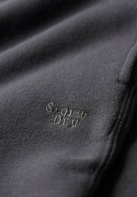Donkergrijs textiel met een soepele textuur, voorzien van een geborduurd "Super Dry"-logo in een lichtere grijstint, prominent gepositioneerd.