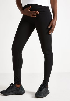 Person iført sorte leggings og sorte sneakers, stående med den ene hånd hvilende på nedre mave og den anden ved siden.