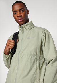 Light green Columbia jacket with a high collar, zippered front, elastic cuffs, and a textured fabric, worn by a person holding a backpack strap.