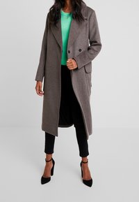 Grau melierter Wollmischung langer Mantel mit doppelter Knopfleiste, Reverskragen, seitlichen Taschen, getragen über einem leuchtend grünen Pullover und schwarzen Hosen.