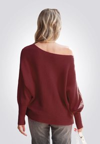 Bordeauxfarbener, übergroßer Strickpullover mit weiter Ausschnitt, gerippten Ärmeln und lockerer Passform, kombiniert mit grauen Jeans und einer weißen Tasche.